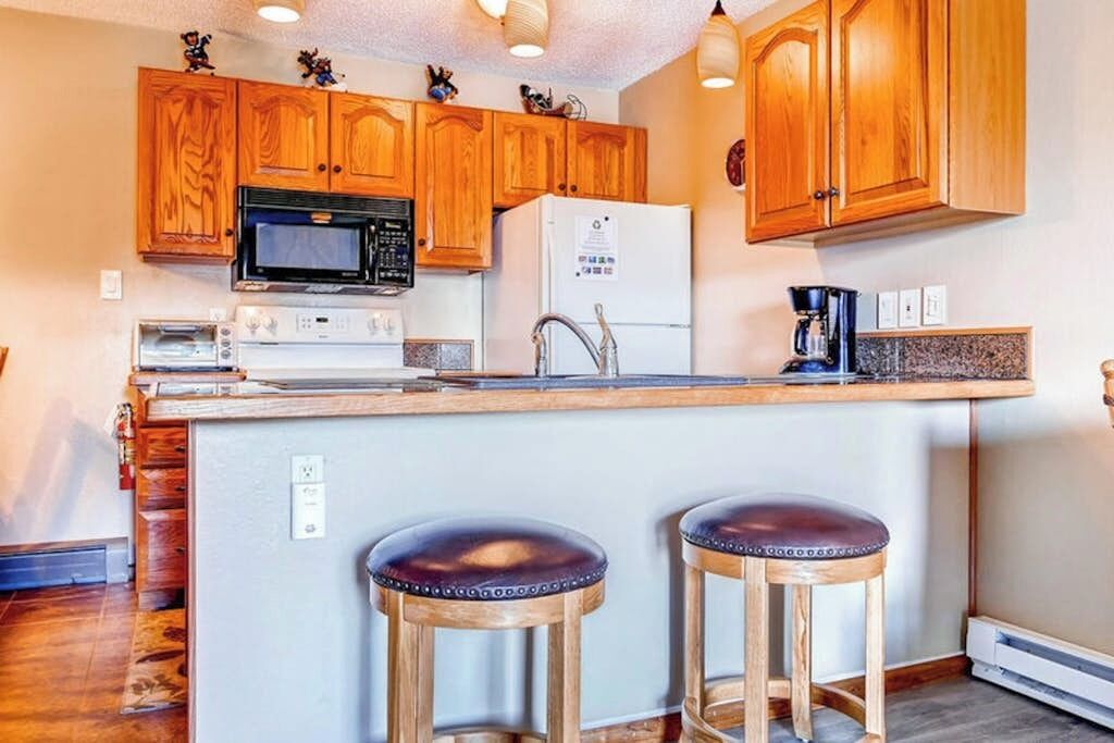 Una cucina con mobili in legno, un forno a microonde, un frigorifero, un tostapane e due sgabelli.