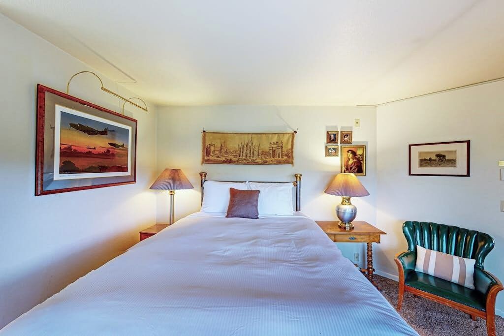 Habitació d'hotel amb un llit gran, dues taules de nit amb làmpades, una cadira tapissada de color verd i obra d'art a les parets.