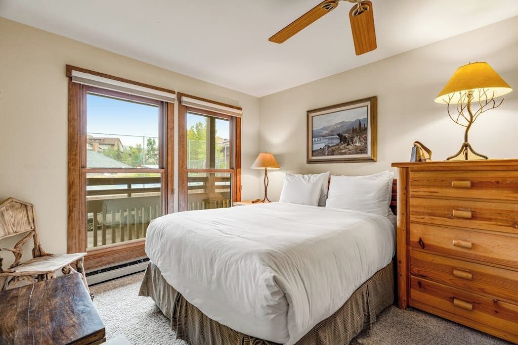 A bedroom with a large bed, a wooden dresser, a desk, a chair, a ceiling fan, and a window with a view.