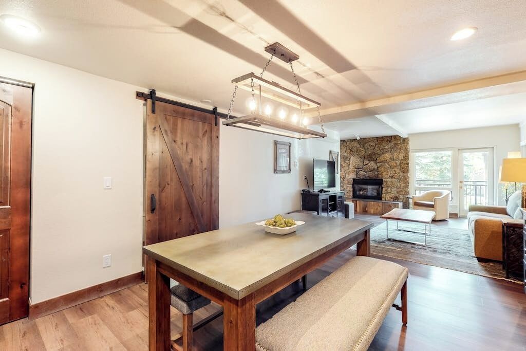 Un vaste espace de vie comprenant une table à manger en bois, un banc, une cheminée en pierre et une grande fenêtre.
