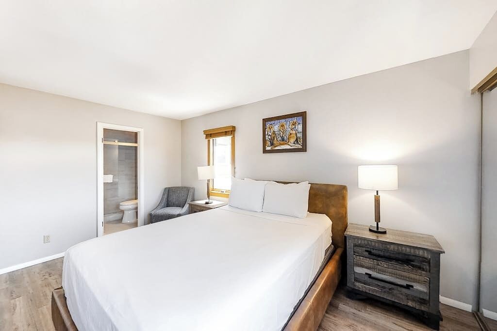 Une chambre à coucher comprenant un lit, une chaise, une table de chevet, un tableau et une porte donnant sur une salle de bain.