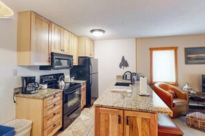 A compact kitchen with wooden cabinets, a microwave, stove, refrigerator, granite countertop, and a small island with a sink. - Great condo w/ remodeled kitchen, balcony, & ski-in/ski-out access to mountain (Steamboat Springs)