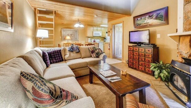 A cozy living room with a sofa, coffee table, fireplace, television, and kitchen in the background. - Ski Inn 231 Lofted family condo with ski-in/ski-out access, trails outside, & shared hot tub (Steamboat Springs)