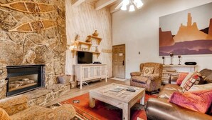 Ein gemütliches Wohnzimmer mit Kamin aus Stein, einem Fernseher und einem großen Gemälde einer Berglandschaft.