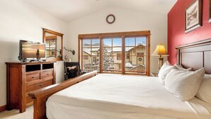 Un dormitorio con cama, cómoda de madera, escritorio, lámpara y ventanas amplias con vista a edificios.