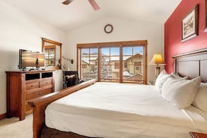 A bedroom with a bed, wooden dresser, desk, lamp, and large windows with a view of buildings. - 1479 EagleRidge Walk-to-lift townhome with office, two decks, mountain views, pool & hot tubs (Steamboat Springs)