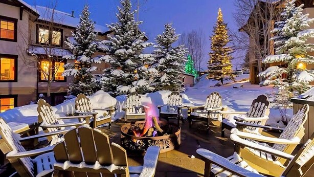 Zona de estar exterior cubierta de nieve con sillas Adirondack y un brasero, rodeada de árboles y edificios cargados de nieve.