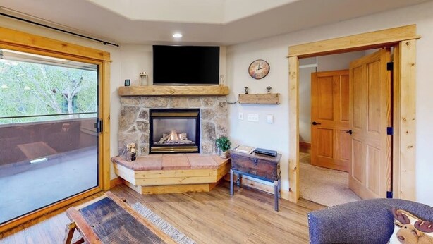 A cozy living room with a stone fireplace, wooden beams, a television, and a large window. - Canyon Creek B306 - Spacious condo with pool, hot tub, gym & easy walk to lifts (Steamboat Springs)