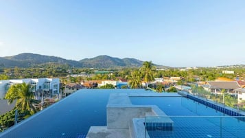 A rooftop pool with solar panels, surrounded by buildings and palm trees, with a view of distant hills.