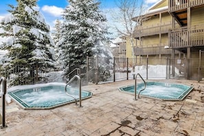 Ein Wellnessbereich mit zwei Whirlpools, einem Holzgebäude und schneebedeckten Bäumen.