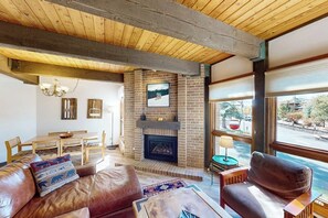 Ein Wohnzimmer mit Kamin, Holzbalken an der Decke, einer Essecke und großen Fenstern.