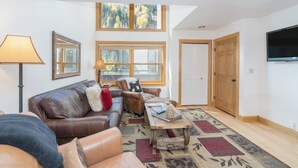 Een woonkamer met een bruine leren bank, een houten salontafel, een lamp en een groot raam met uitzicht op bomen.