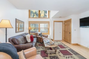 A living room with a brown leather couch, a wooden coffee table, a lamp, and a large window with a view of trees. - Ski-in/out condo with hot tub, grill, balcony & covered parking (Telluride)