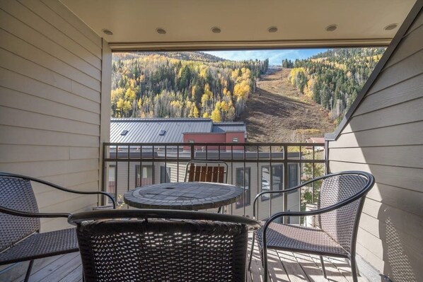 A balcony with wicker chairs and a small table, overlooking a forested hillside. - Ski-in/out condo with hot tub, grill, balcony & covered parking (Telluride)