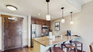 A kitchen with a granite countertop, wooden stools, a refrigerator, a microwave, and pendant lighting. - Convenient Home Close to Steamboat Ski Resort, Hiking Trails, Restaurants & More (Steamboat Springs)