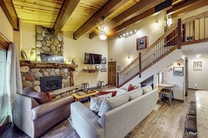 Ein geräumiges Wohnzimmer mit Kamin aus Naturstein, einer großen Couch, einem Fernseher und einer Treppe, die zu einer oberen Ebene führt.