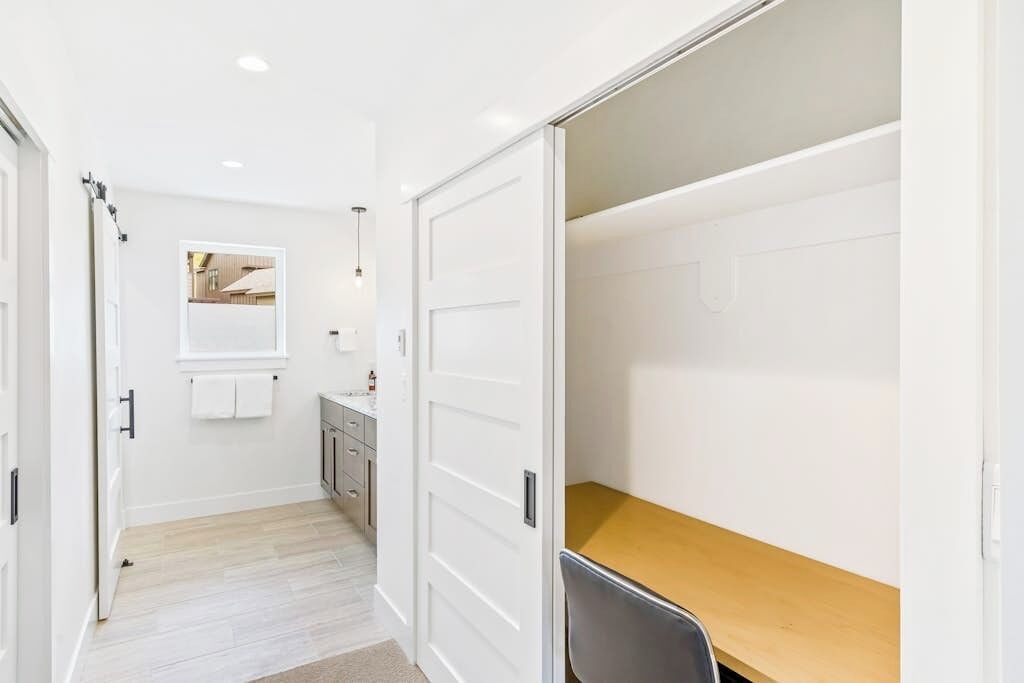 Un pasillo con suelo de madera, una puerta blanca, un espejo, un lavamanos pequeño y una mesa de madera.