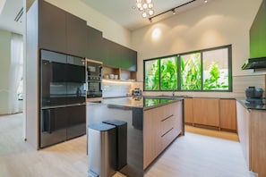 A modern kitchen with a central island, built-in appliances, and a large window with a view of greenery. - Ruby Luxury Villa, 4 Bed Private Pool Paradise (Rawai)