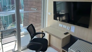 A modern hotel room with a large TV, a desk, a chair, and a view of the outside balcony.
