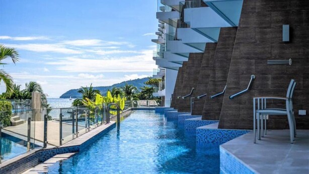 A modern hotel with a long swimming pool, a view of the sea, and a mountain in the background.
