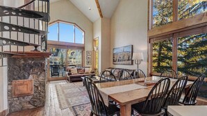 A spacious room with a dining table, chairs, a fireplace, a staircase, and large windows with a view of trees. - Fantastic, ski-in/out condo with patio, fireplace, grill & Washer/Dryer (Mountain Village)
