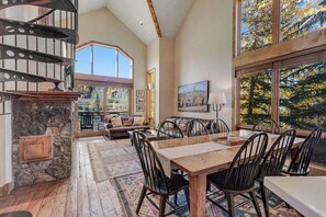A spacious room with a dining table, chairs, a fireplace, a staircase, and large windows with a view of trees. - Fantastic, ski-in/out condo with patio, fireplace, grill & Washer/Dryer (Mountain Village)