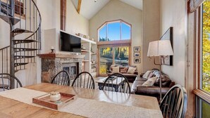 A spacious living area with a fireplace, a dining table, a sofa, and a large window offering a view of trees. - Fantastic, ski-in/out condo with patio, fireplace, grill & Washer/Dryer (Mountain Village)