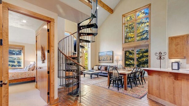 A spacious room with a spiral staircase, a dining area with chairs, a sofa, and a large window with a view of trees. - Fantastic, ski-in/out condo with patio, fireplace, grill & Washer/Dryer (Mountain Village)