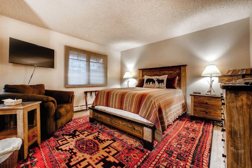 A bedroom with a bed, a chair, a TV, a window with blinds, and a patterned rug.