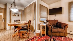 Een kamer met een houten eettafel, stoelen, een bruine leren bank, een aan de muur bevestigde televisie en een kamerkleed met een patroon.