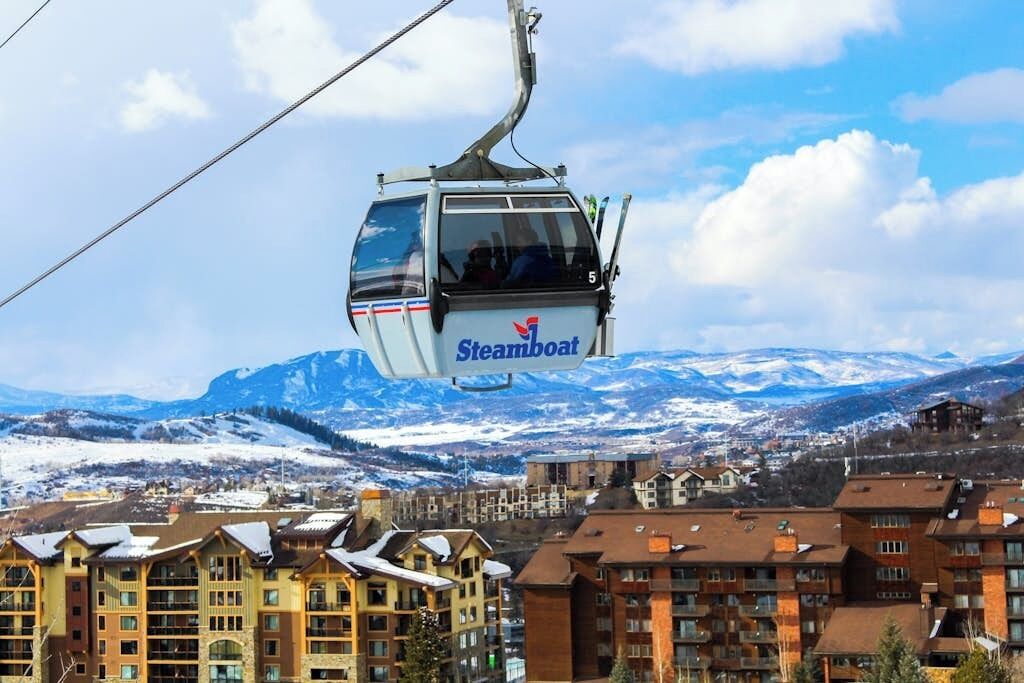 Un teleférico con la marca 'Steamboar', un telón de fondo montañoso y una ciudad con edificios de varios pisos.