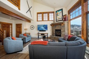 A spacious living room with a stone fireplace, large windows, and a television mounted on the wall. - Spacious Corner Home w/ Incredible View, Free WiFi, Private Hot Tub & Large Deck (Steamboat Springs)