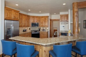 A kitchen with wooden cabinets, granite countertops, a stainless steel refrigerator, a microwave, a stove, and blue bar stools. - Spacious Corner Home w/ Incredible View, Free WiFi, Private Hot Tub & Large Deck (Steamboat Springs)