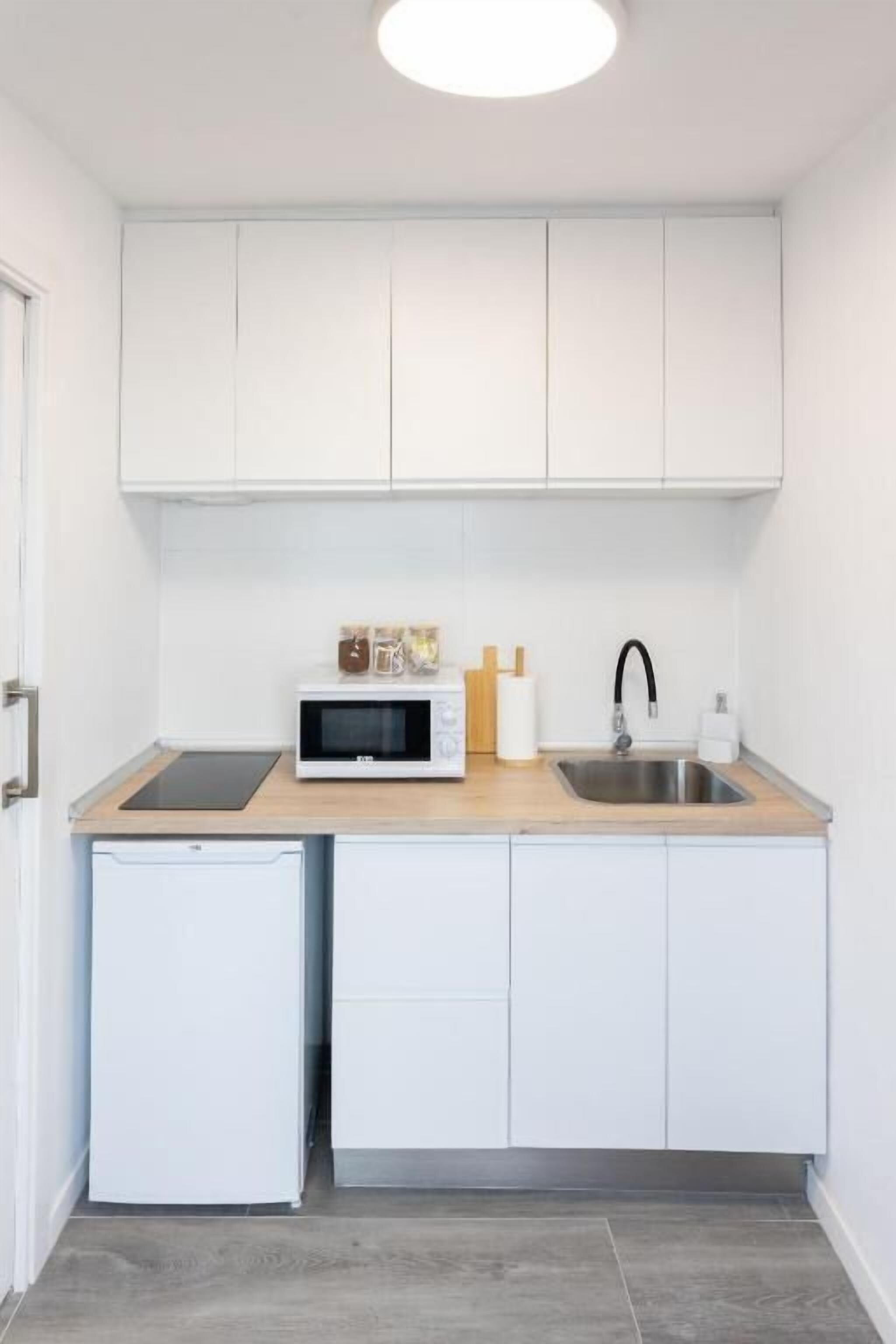 Een kleine keuken met witte kastjes, een magnetron, een spoelbak en een kleine koelkast.