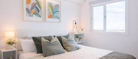 A bedroom with a bed, bedside tables, wall art, a window, and an air conditioning unit.