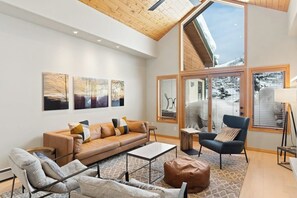 Ein geräumiges Wohnzimmer mit hoher Decke, einem großen Fenster mit Jalousien, einer Couch, Sesseln und Kunstwerken an der Wand.