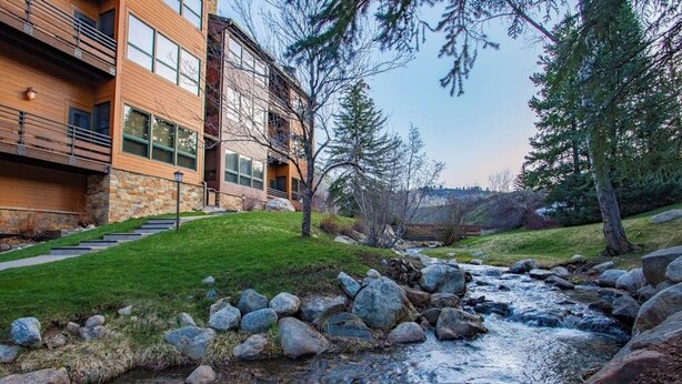 A modern building with a stone facade, a grassy area, and a stream. - Kutuk 101 - First-Floor Condo with Shared Hot Tub, WiFi, Gas Fireplace, and Full Kitchen (Steamboat Springs)