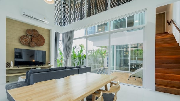 A modern living room with a wooden dining table, grey sofas, a flat-screen TV, and large windows overlooking a garden. - NEW Lavender Private Pool Villa, Rawai , Phuket (Nai harn, Rawai)