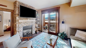 Ein Wohnzimmer mit Kamin aus Stein, einem Flachbildfernseher, einer Couch, einem Couchtisch und Blick durch große Glasüren.