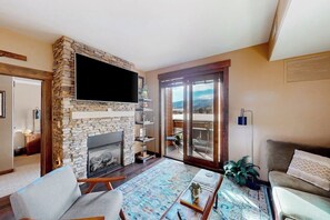 Ein Wohnzimmer mit Kamin aus Stein, einem Flachbildfernseher, einer Couch, einem Couchtisch und Blick durch große Glasüren.