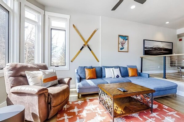 A living room with a brown leather armchair, a blue sofa with orange cushions, a wooden coffee table, and a red patterned rug. - Ski view condo walkable to the lifts with pools, hot tubs, WiFi, and fireplace (Steamboat Springs)