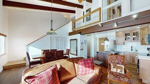 A spacious living area with a high ceiling, a dining table with chairs, a kitchen with wooden cabinets, and a staircase leading to an upper level. - Two-Story Ski-In Condo w/Free WiFi, Gas Fireplace, Private W/D, Shared Hot Tub (Steamboat Springs)
