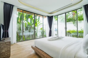 Ein modernes Schlafzimmer mit einem großen Bett, einem Fernseher und Blick auf üppiges Grün durch raumhohe Fenster.
