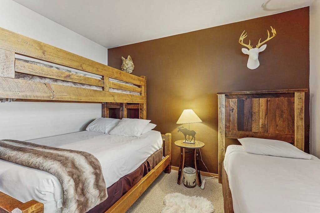 A room with a wooden bed, a small table with a lamp, and a wall-mounted deer head.