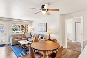 A living room with a round wooden table, two sofas, a ceiling fan, and a large window. - Renovated, ground-floor condo with pool, hot tubs & gym - steps to gondola (Steamboat Springs)