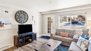 Ein Wohnzimmer mit einer großen Uhr, einem Fernseher, einem Sofa und Blick ins Freie.
