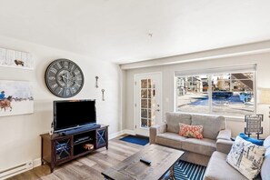 A living room with a large clock, a television, a sofa, and a view of the outside. - Renovated, ground-floor condo with pool, hot tubs & gym - steps to gondola (Steamboat Springs)