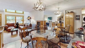 Un salón espacioso con una mesa redonda de cristal, sillas de madera y una araña. Se ve una cocina al fondo con armarios de madera y una zona de comedor.