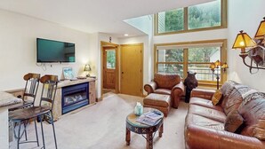 Un amplio salón con chimenea, muebles de cuero, televisor y ventanas grandes.