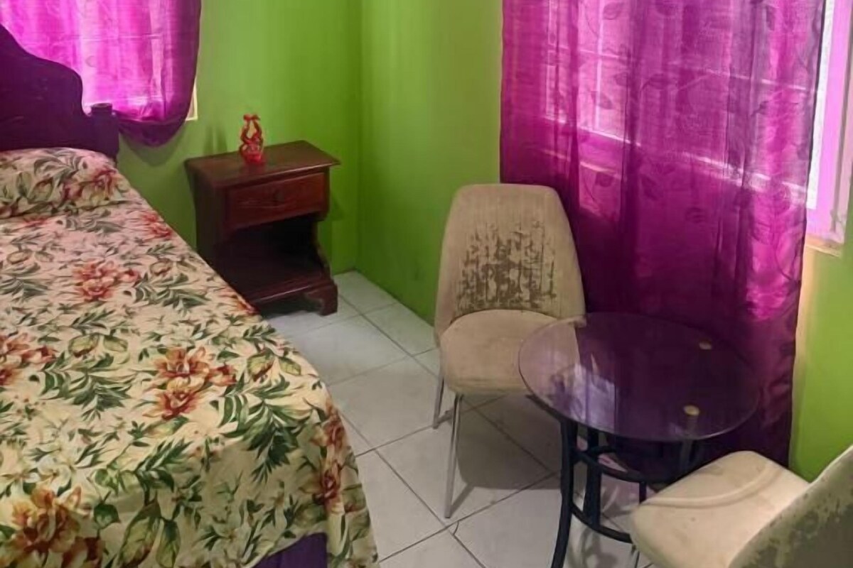A room with a bed, a chair, a small table, an air conditioner, and a window with purple curtains.